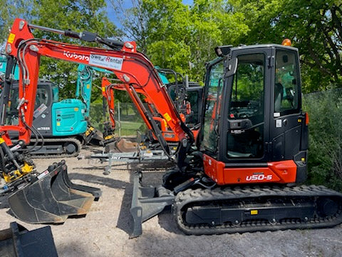 Kompakt bandgrävare utrustad med rototilt och grip samt tre skopor, uppställd på arbetsplats med fler maskiner bakom